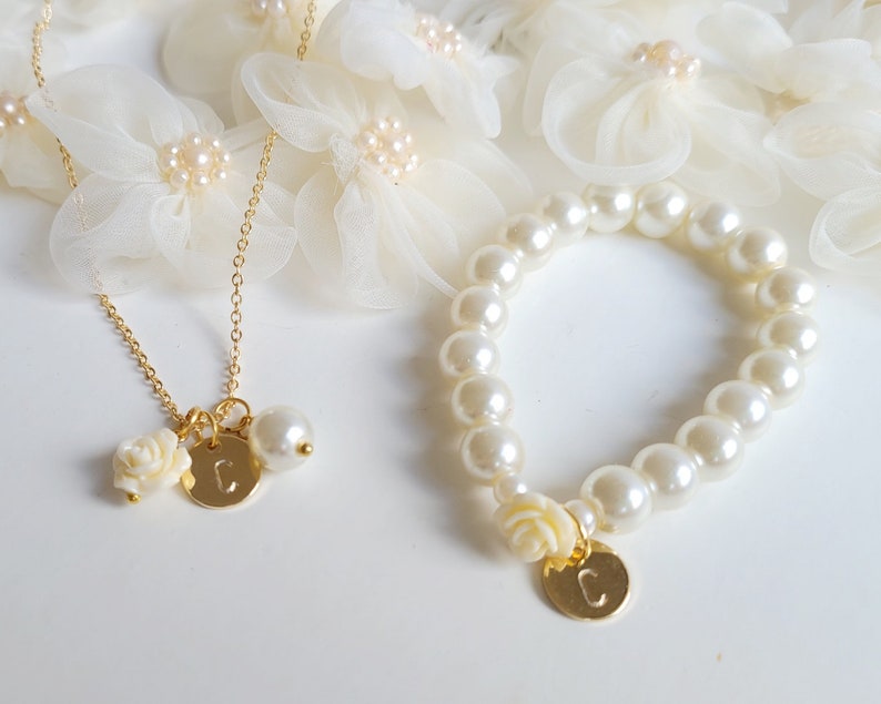 K&ouml;nnte beinhalten: Ein goldenes Halsketten- und Armband-Set mit Perlenakzenten und einem Anh&auml;nger mit dem Buchstaben "C". Die Anh&auml;nger sind eine kleine wei&szlig;e Blume und eine runde Scheibe mit dem Buchstaben "C".