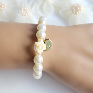 K&ouml;nnte beinhalten: Ein wei&szlig;es Perlenarmband mit einem goldenen Anh&auml;nger, der eine wei&szlig;e Rose und den Buchstaben "S" zeigt.