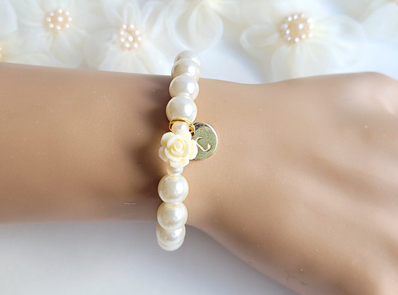 K&ouml;nnte beinhalten: Ein wei&szlig;es Perlenarmband mit einem goldenen Anh&auml;nger, der eine wei&szlig;e Rose und den Buchstaben "S" zeigt.