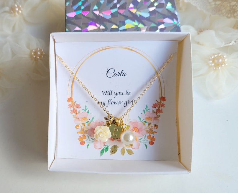 K&ouml;nnte beinhalten: Eine wei&szlig;e Schachtel mit einer goldenen Halskette darin. Die Halskette hat einen kleinen Blumenanh&auml;nger und einen Perlenanh&auml;nger. Die Schachtel hat eine Karte mit der Aufschrift "Carla, Willst du meine Blumenm&auml;dchen sein?"