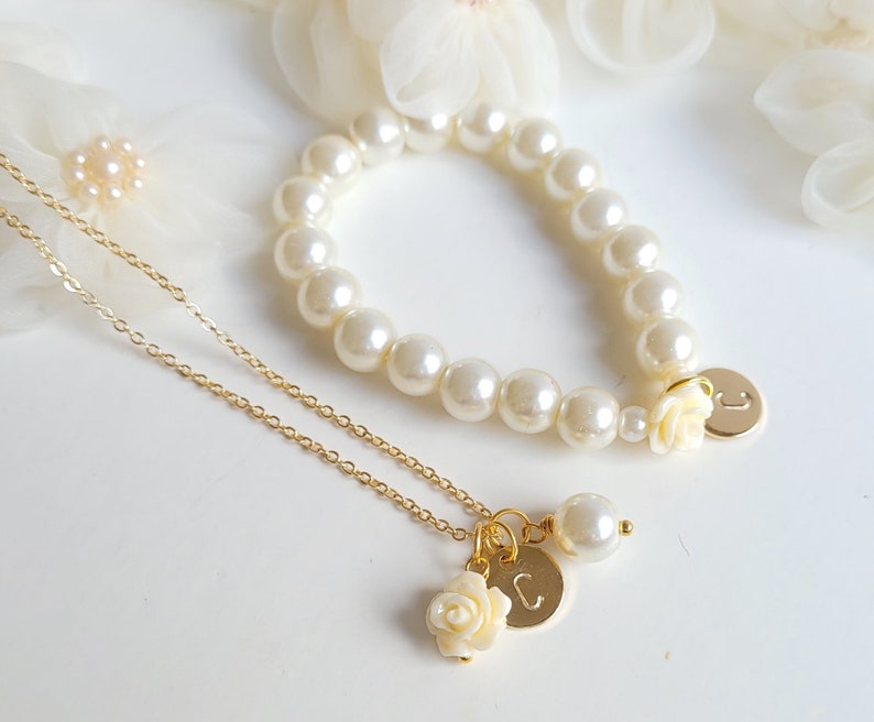 K&ouml;nnte beinhalten: Eine goldene Kettenkette mit einem kleinen wei&szlig;en Rosenanh&auml;nger und einem goldenen Initialanh&auml;nger. Ein Perlenarmband mit einem wei&szlig;en Rosenanh&auml;nger und einem goldenen Initialanh&auml;nger.