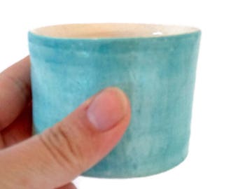 May include: A small, handmade ceramic cup with a light blue exterior and a beige interior. The cup has a cylindrical shape and a slightly textured surface. The cup is held by a hand against a white background.
