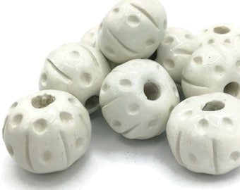 Peut inclure: Un ensemble de perles en céramique blanc cassé. Chaque perle sphérique présente un trou central et de petits trous décoratifs ainsi que des lignes sculptées. Les perles sont regroupées sur un fond blanc, mettant en valeur leur texture et leur design uniques. Mesures en centimètres.
