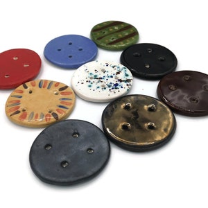 Könnte beinhalten: Eine Sammlung handgefertigter Keramikknöpfe in verschiedenen Farben und Designs. Die Knöpfe sind rund und oval, mit mehreren Löchern zum Annähen. Farben sind Rot, Blau, Schwarz, Braun und Weiß mit dekorativen Mustern.