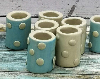 Puede incluir: Colección de jarrones cilíndricos de cerámica en azul claro y crema. Cada jarrón está adornado con lunares en relieve de color crema. Los jarrones tienen un acabado liso y brillante y están colocados sobre una superficie de madera envejecida y multicolor.