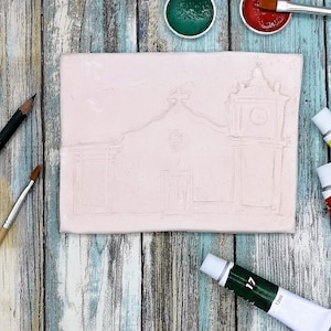 Op de afbeelding: Een lichtroze rechthoekige keramische tegel met een geëtst architectonisch ontwerp van een gebouw met een klokkentoren. Verftubes, penselen en verfpotten in rood en groen zijn rond de tegel op een houten oppervlak geplaatst.