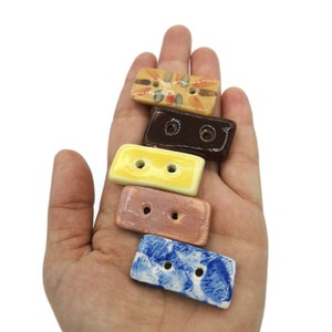 May include: A collection of five rectangular ceramic buttons in various colors and patterns. The buttons are held in a hand and feature two holes each for sewing. Colors include yellow, brown, blue, and a tan with a floral design.