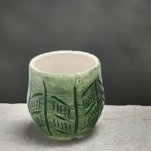 Puede incluir: Un pequeño macetero de cerámica verde con un patrón geométrico. El macetero está sobre una superficie gris claro.