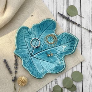 Op de afbeelding: Een blauwgroen keramieken schaaltje in de vorm van een esdoornblad met een glanzende afwerking en zichtbare nerven. Het schaaltje bevat een zilveren ring met een donkere steen, een fijne gouden ketting en een paar kleine gouden oorknopjes.