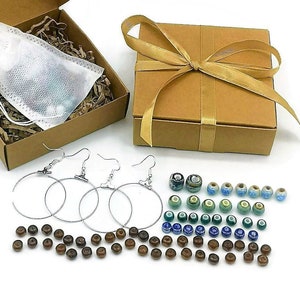 May include: A craft kit with a variety of jewelry-making supplies. Includes a box with a white pouch, a closed box with a gold ribbon, silver hoop earrings, wooden beads, and colorful glass beads. The kit is presented on a white background.