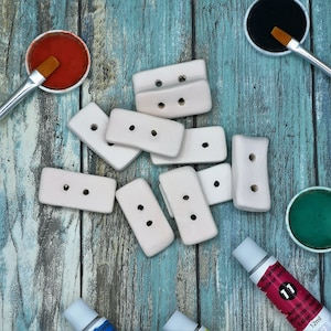 May include: A collection of rectangular, off-white wooden buttons with two holes each, scattered on a weathered, turquoise-painted wooden surface. Paint pots with brushes and acrylic paint tubes in various colors are also present, suggesting a crafting or art theme.