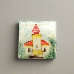 Puede incluir: Un azulejo de cerámica pintado a mano que representa un faro con un techo rojo, paredes amarillas y molduras blancas. El faro se encuentra sobre un fondo de acuarela azul y blanco con árboles verdes a cada lado.
