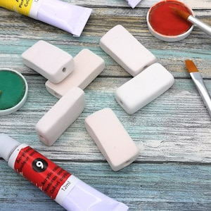 May include: A collection of rectangular, off-white beads with a hole drilled through the center, arranged on a weathered, blue-green wooden surface. Paint tubes, paintbrushes, and paint pots are also visible, suggesting an art or craft theme.