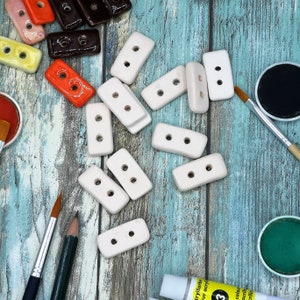 Op de afbeelding: Overzicht van diverse keramische knopen in meerdere kleuren, waaronder wit, oranje, geel en bruin. De knopen zijn rechthoekig met elk twee gaten. Penselen, verf en potloden liggen verspreid rond de knopen op een blauw-witte houten ondergrond.
