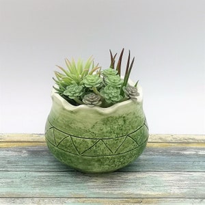 May include: A round, green ceramic planter with a scalloped rim and a horizontal band of incised triangles. The planter is filled with a variety of artificial succulents in shades of light green, sage, and dark reddish-brown, creating a dense arrangement.