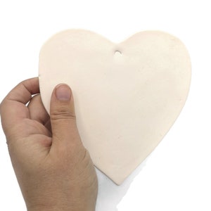 May include: A heart-shaped, off-white ceramic ornament with a small hole at the top for hanging. The ornament is held in a hand against a white background. The heart is smooth and has a matte finish.