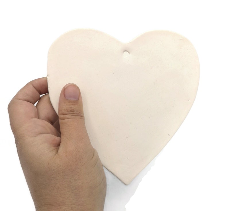 May include: A heart-shaped, off-white ceramic ornament with a small hole at the top for hanging. The ornament is held in a hand against a white background. The heart is smooth and has a matte finish.