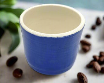 May include: A blue ceramic coffee cup with a cream-colored interior and a white rim. The cup is surrounded by scattered coffee beans. The cup is a small size, perfect for espresso or a small coffee.