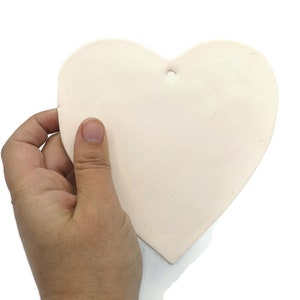 May include: A light pink heart-shaped ceramic ornament with a small hole at the top for hanging. The ornament is held in a hand against a white background. The heart is smooth and has a matte finish.