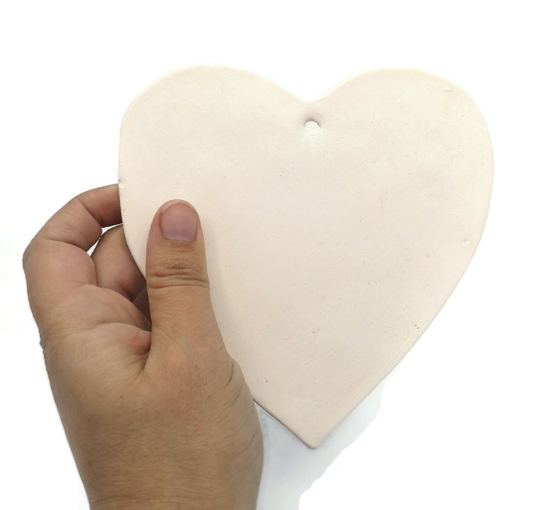 May include: A light pink heart-shaped ceramic ornament with a small hole at the top for hanging. The ornament is held in a hand against a white background. The heart is smooth and has a matte finish.