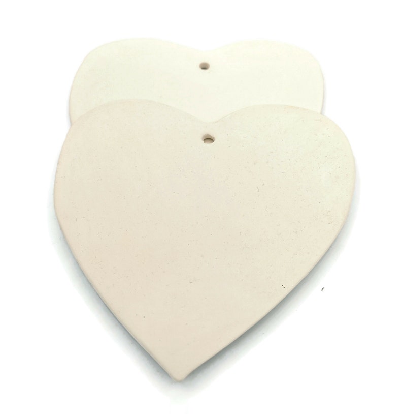 May include: Two off-white, heart-shaped ornaments with a textured surface. Each heart has a small hole at the top for hanging. The hearts are stacked slightly, with one in front of the other, against a white background.