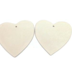 May include: Two off-white heart-shaped ornaments with a matte finish. Each heart has a small hole at the top for hanging. The hearts are displayed against a white background.