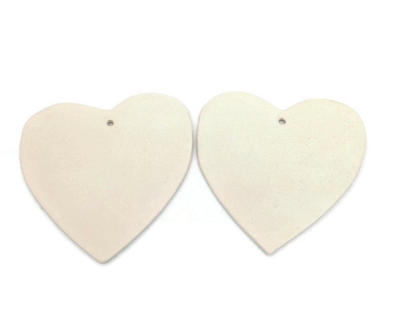 May include: Two off-white heart-shaped ornaments with a matte finish. Each heart has a small hole at the top for hanging. The hearts are displayed against a white background.