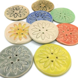 Puede incluir: Nueve botones de cerámica con un diseño de margarita en varios colores, incluyendo amarillo, azul, verde, naranja y blanco.