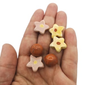 May include: A collection of ceramic flower and sphere shaped beads in shades of yellow, orange, and pink. The beads are arranged on a hand.