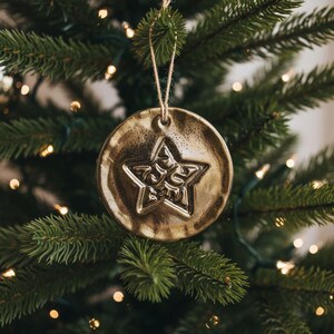 Op de afbeelding: Een rond, goudkleurig ornament met een textuurrijk, onregelmatig oppervlak. In het midden is een vijfpuntige ster aangebracht met ingewikkelde, wervelende patronen. Het ornament hangt aan een dunne, lichtgekleurde lus van touw.