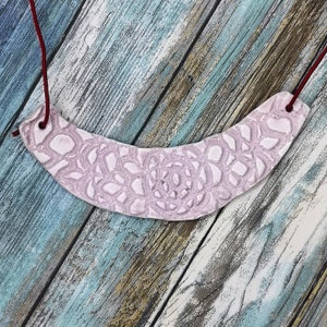 May include: A pink ceramic necklace with a delicate lace pattern. The necklace is strung on a red cord.