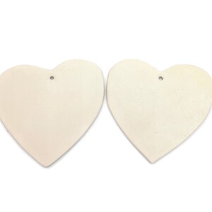 May include: Two off-white heart-shaped ornaments with a matte finish. Each heart has a small hole at the top for hanging. The hearts are displayed against a white background, highlighting their simple, elegant design.