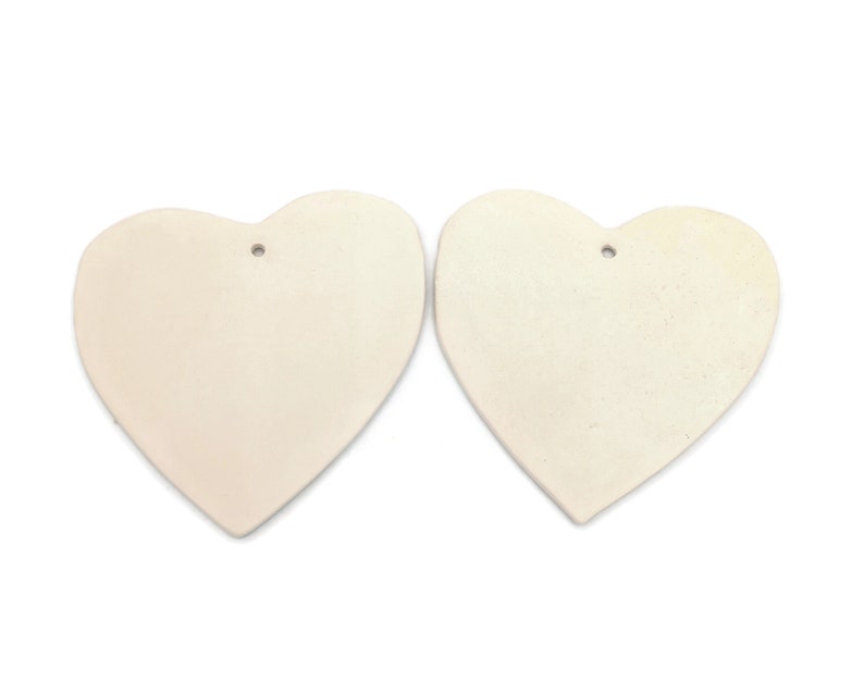 May include: Two off-white heart-shaped ornaments with a matte finish. Each heart has a small hole at the top for hanging. The hearts are displayed against a white background, highlighting their simple, elegant design.