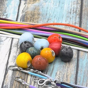 Può includere: Una collezione di perline in ceramica colorate di varie forme e dimensioni, tra cui gialle, rosse e blu. Sono visibili diversi cordini di diversi colori, insieme a chiusure, che suggeriscono forniture per la creazione di gioielli. Le perline sono disposte su una superficie di legno chiaro.