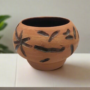 May include: A terracotta ceramic pot with a dark interior and a rounded shape. The exterior features dark brown abstract designs. The pot sits on a white surface and is suitable for plants or decorative use.