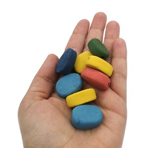 May include: A hand holding a collection of colorful, smooth, stone-like beads. The beads are in shades of blue, yellow, red, and green. Each bead has a hole, suggesting they are for stringing into jewelry or other crafts.