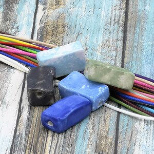 Puede incluir: Surtido de cuentas de cerámica de colores con agujeros para enhebrar. Las cuentas son rectangulares y tienen un acabado esmaltado. Los colores incluyen azul, verde, negro y blanco.