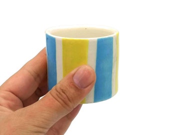 May include: A small, white ceramic cup with blue and yellow vertical stripes. The cup is being held in a hand.