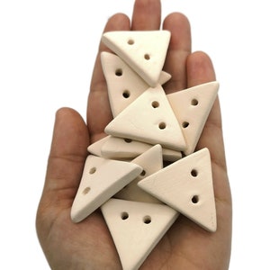 Puede incluir: Un puñado de botones triangulares de color blanquecino, cada uno con dos agujeros. Los botones son de un material mate y tienen una forma uniforme. Los botones se sostienen en una mano abierta sobre un fondo blanco.