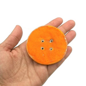 Puede incluir: Un botón grande y redondo de color naranja con cuatro agujeros, sostenido en una mano. El botón tiene una superficie texturizada y un borde ligeramente blanquecino. El botón es de color naranja brillante.