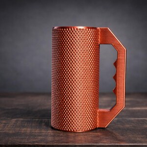 Peut inclure: Une tasse cylindrique de couleur cuivre avec une surface ext&eacute;rieure textur&eacute;e &agrave; motif de losanges. La tasse comprend une poign&eacute;e g&eacute;om&eacute;trique avec trois empreintes digitales distinctes sur le bord int&eacute;rieur pour une prise en main.