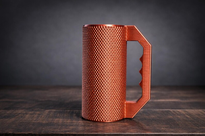 Peut inclure: Une tasse cylindrique de couleur cuivre avec une surface ext&eacute;rieure textur&eacute;e &agrave; motif de losanges. La tasse comprend une poign&eacute;e g&eacute;om&eacute;trique avec trois empreintes digitales distinctes sur le bord int&eacute;rieur pour une prise en main.