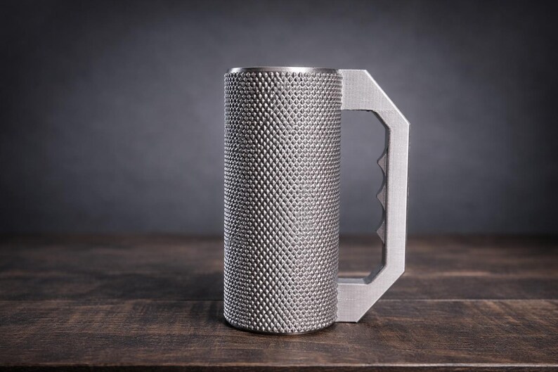 Peut inclure: Une tasse cylindrique en m&eacute;tal avec une surface textur&eacute;e &agrave; motif de losanges et une poign&eacute;e plate et angulaire comportant une s&eacute;rie de rainures pour les doigts sur le c&ocirc;t&eacute; int&eacute;rieur. La tasse est faite d'un mat&eacute;riau argent&eacute; mat.