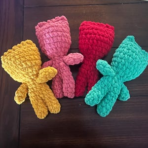 May include: Four handmade crochet dolls in yellow, pink, red, and teal. Each doll has a rounded head, small arms, and legs. The dolls are made with a soft, textured yarn.