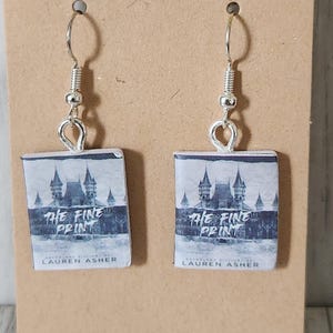 May include: A pair of silver-toned dangle earrings. Each earring features a small, rectangular charm with a book cover design. The book cover displays a castle illustration and the title "THE FINE PRINT" by Lauren Asher.