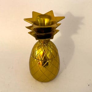 May include: A gold-coloured pineapple-shaped container with a textured surface. The top of the container has a starburst design. The container is likely used for decorative purposes or as a small storage item.