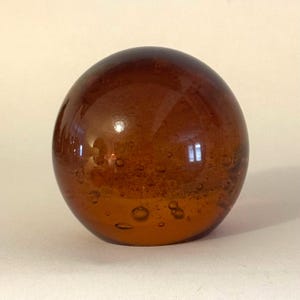 Puede incluir: Una esfera de vidrio transparente de color ámbar con burbujas de aire en su interior. El objeto esférico tiene una superficie brillante que refleja la luz. La bola de cristal es un objeto decorativo, posiblemente un pisapapeles o un adorno.
