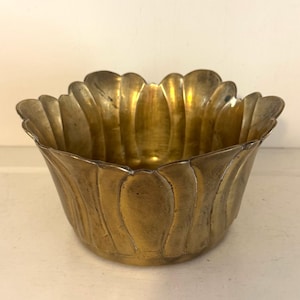 May include: A brass bowl with a scalloped, petal-like rim and a rounded base. The metal surface features a warm, aged golden tone with subtle variations in color and texture throughout the exterior and interior of the vessel.