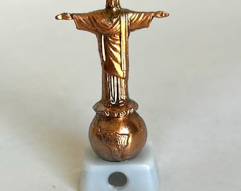 May include: A bronze-colored miniature statue of Christ the Redeemer with outstretched arms, atop a globe-shaped base. The statue is mounted on a white rectangular base with a hole. The statue is a detailed replica of the iconic landmark.