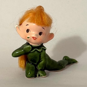 Könnte beinhalten: Keramikfigur eines sitzenden Elfen mit orangefarbenem Haar und einem grünen Outfit. Der Elf hat ein lächelndes Gesicht mit großen Augen und spitzen Ohren. Die Figur hat eine glänzende Oberfläche und ist wahrscheinlich vintage.